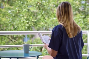 Una persona sentada a una mesa con un libro y una taza, con vista a un balcón con follaje.