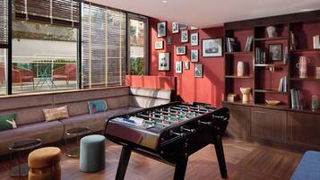 Ein gemütliches Zimmer mit einem Kicker, einer Couch mit Kissen, einem Bücherregal und einem Fenster mit Jalousien.
