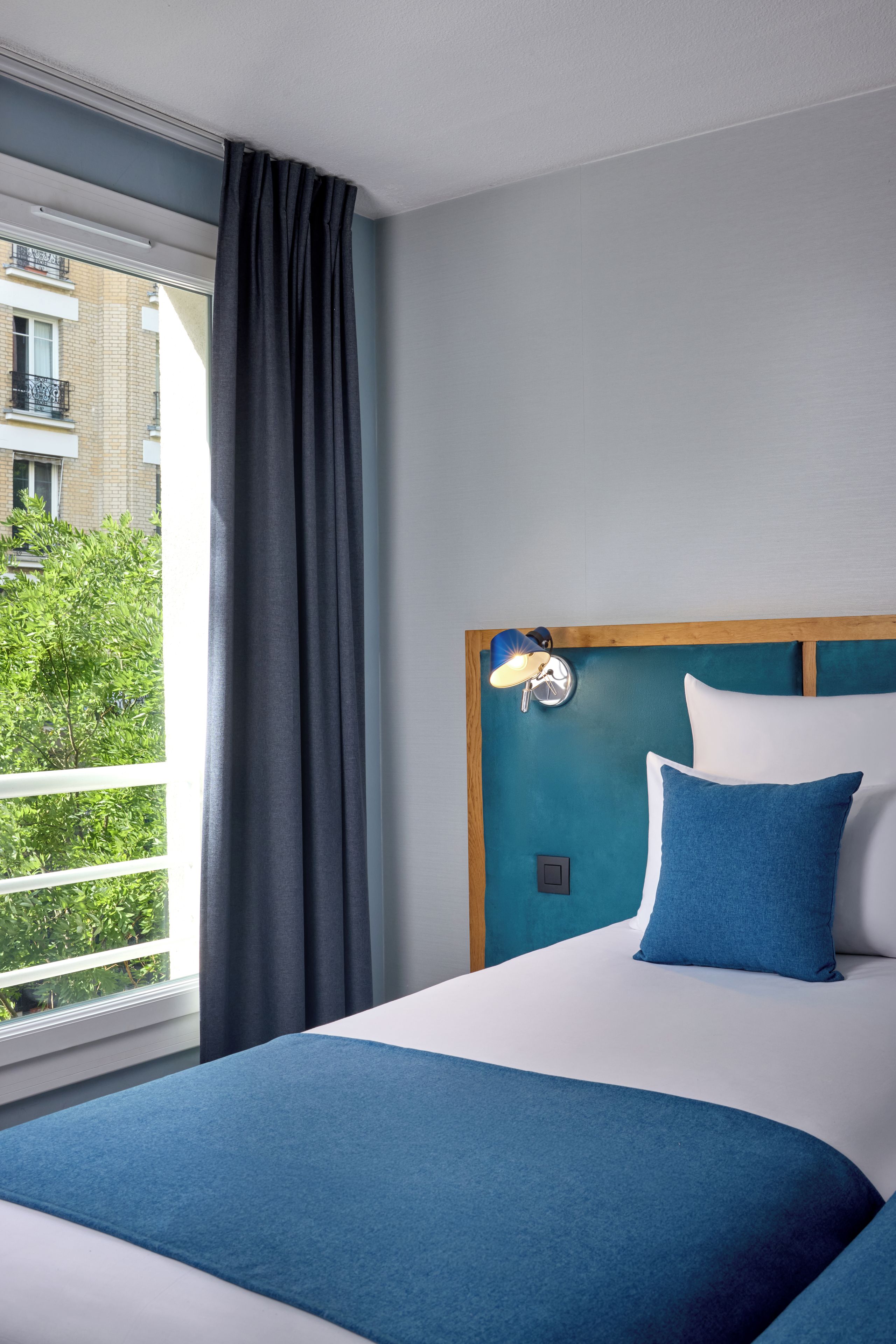 A hotel room with a bed, a window with curtains, a view of greenery outside, and a wall-mounted light fixture.
