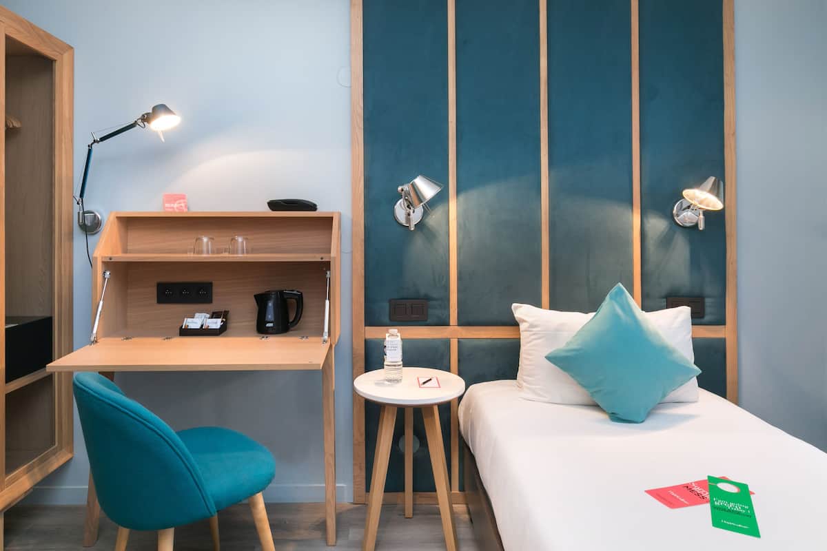 Une chambre d’hôtel avec un bureau en bois, une chaise de couleur turquoise, un lit avec une literie blanche et un oreiller turquoise, une petite table ronde et deux lampes fixées au mur.