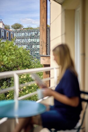 A person seated at a round table on a balcony with a view of a modern building and greenery. - Hôtel Sanso by HappyCulture (Ex Quality Bercy Bibliotèque) (Paris)