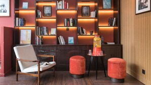 Un coin lecture moderne, doté d’un fauteuil confortable, de deux poufs, d’une petite table avec un distributeur de boissons et d’étagères remplies de divers livres et de photos encadrées.