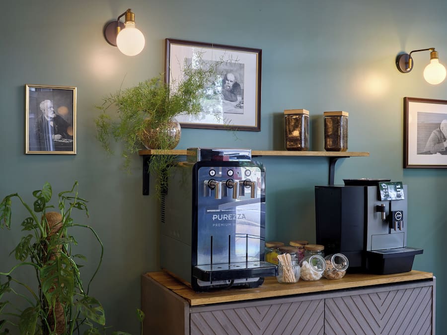 A coffee bar with a Purozza espresso machine, two wall-mounted coffee dispensers, and framed artwork on the wall.