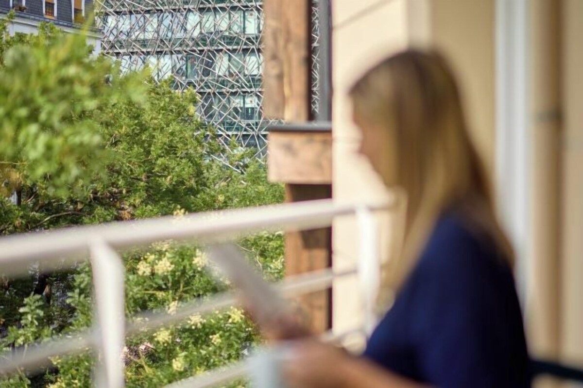 A person standing on a balcony with a railing, overlooking a building with a metal framework and greenery.