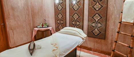A massage therapy room with a massage table, a small table with bottles and a flower, and a decorative wall panel.