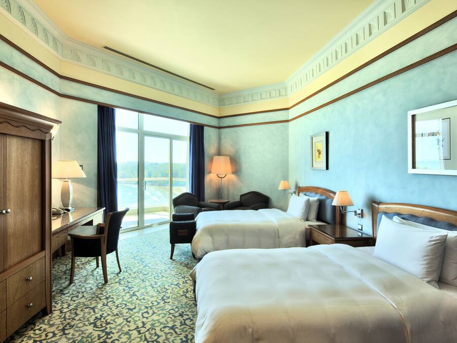 A hotel room with two beds, a desk, a chair, and a large window.