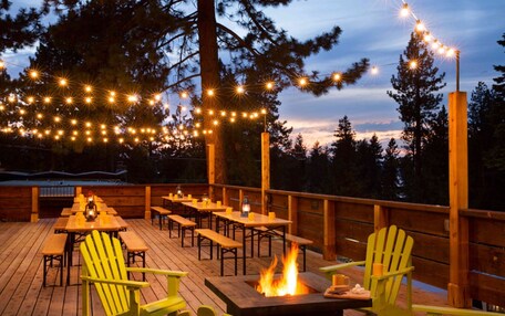 An outdoor deck with wooden tables and chairs, a fire pit, and string lights.