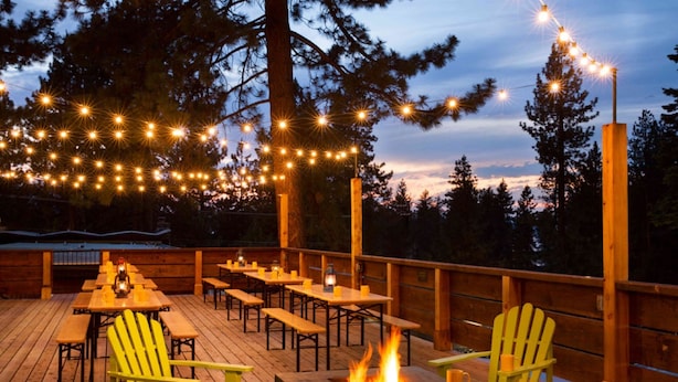Una terraza al aire libre con mesas y sillas de madera, una chimenea y luces colgantes.
