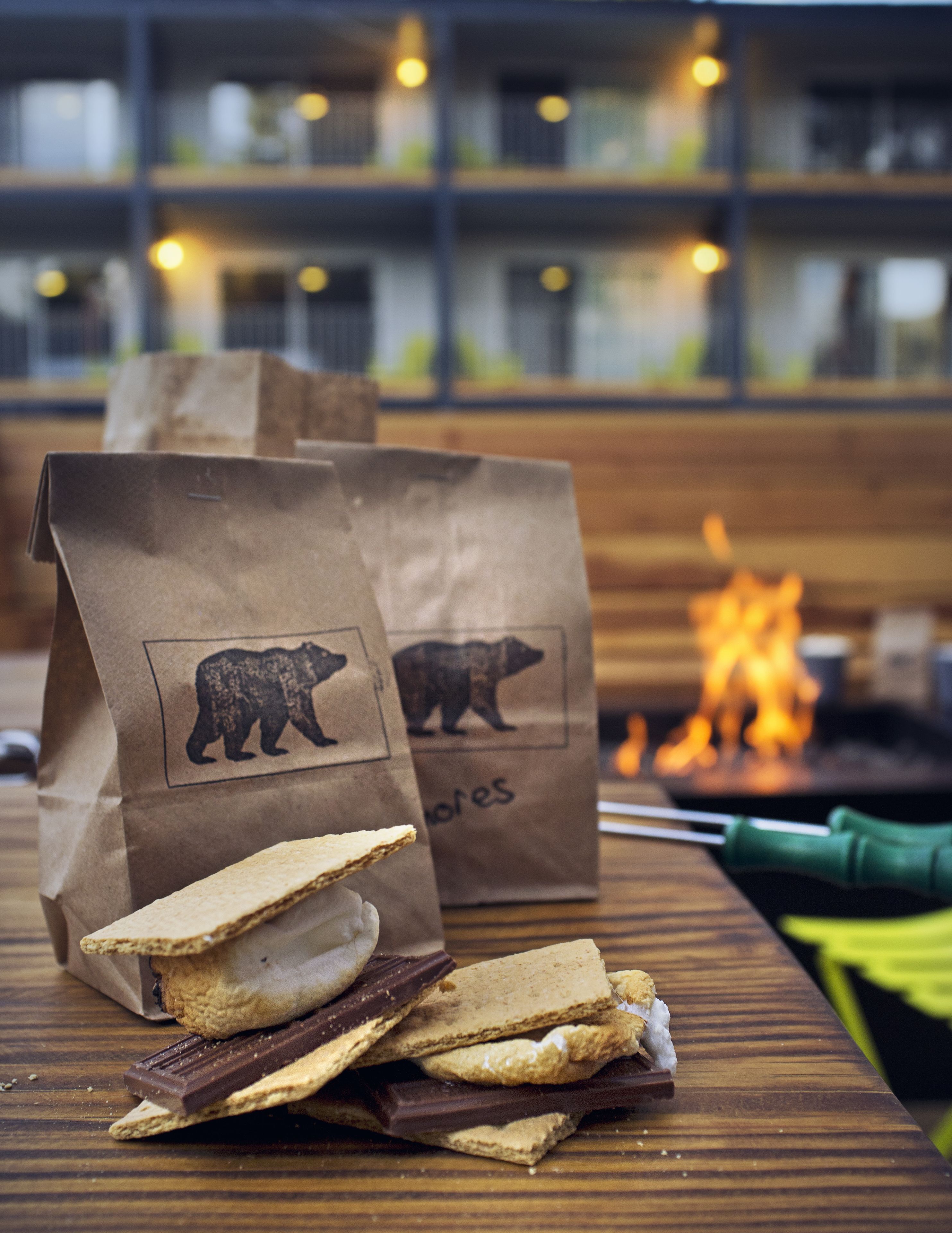 A wooden table with a stack of graham crackers and chocolate bars, two brown paper bags with bear logos, and a green-handled knife.