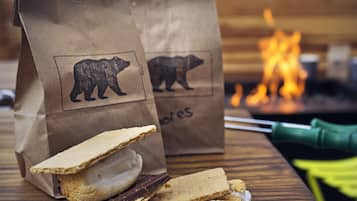 A wooden table with a stack of graham crackers and chocolate bars, two brown paper bags with bear logos, and a green-handled knife.