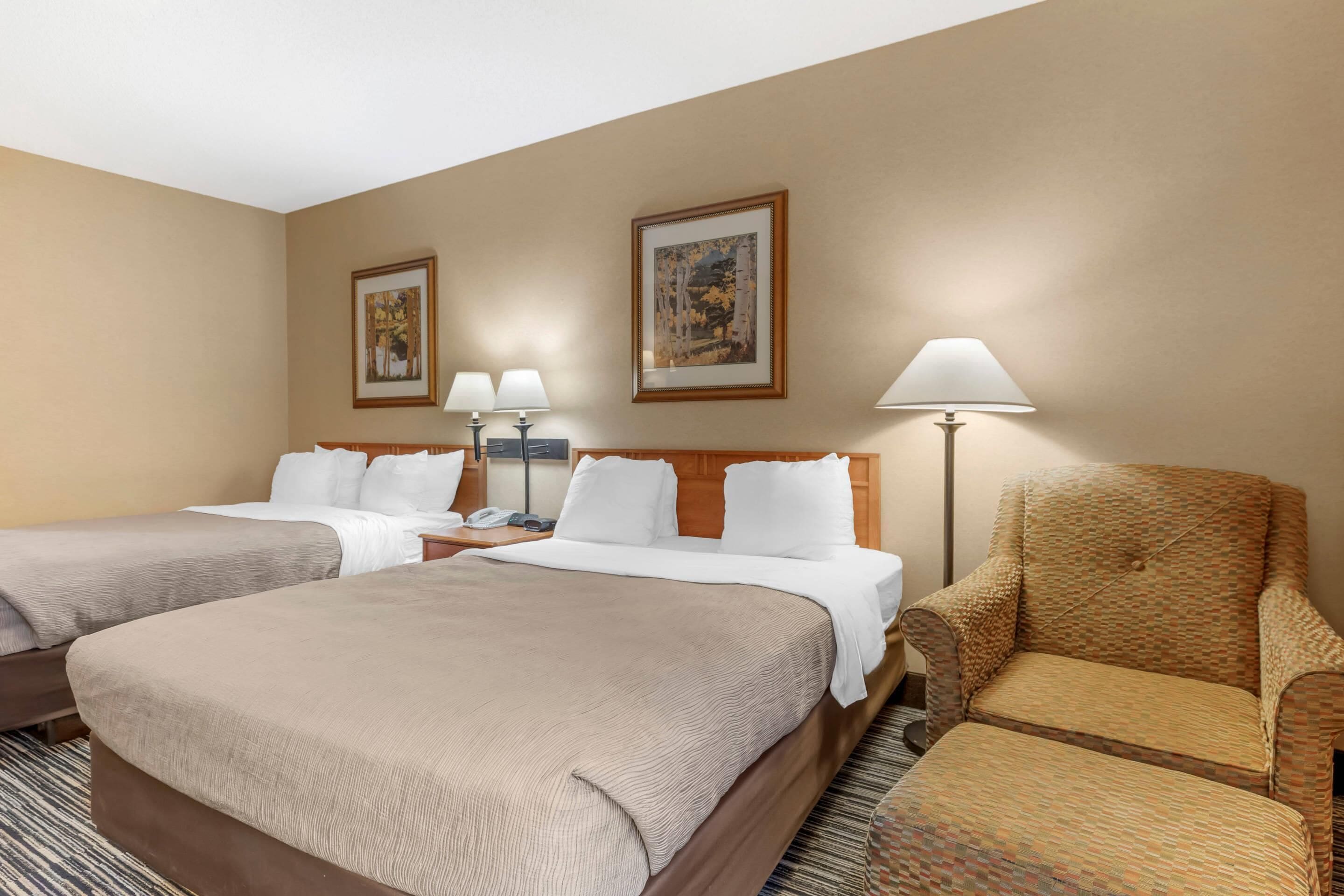 A hotel room with two beds, a chair, a lamp, and two framed pictures on the wall.