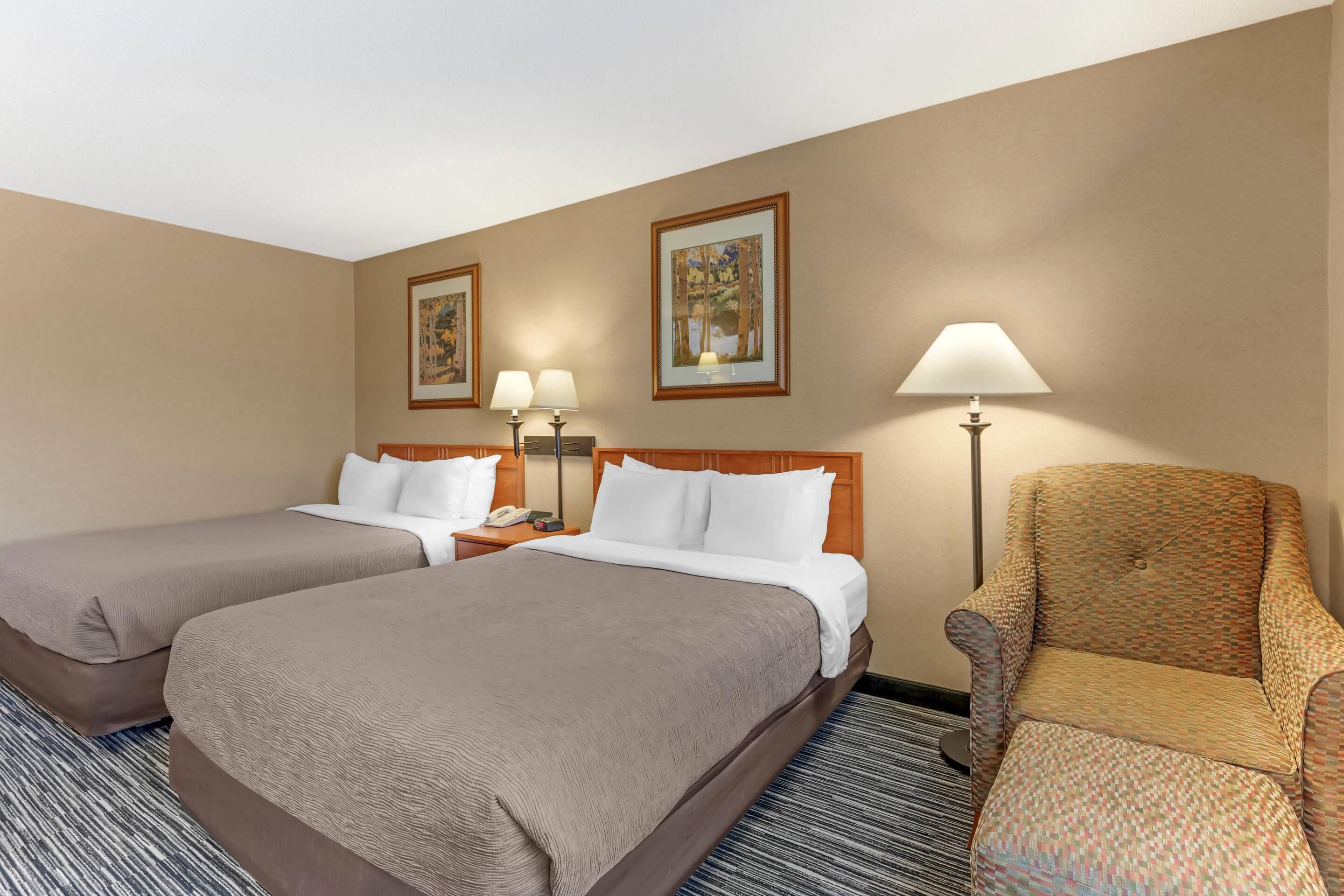 A hotel room with two beds, a chair, a lamp, and framed pictures on the wall.