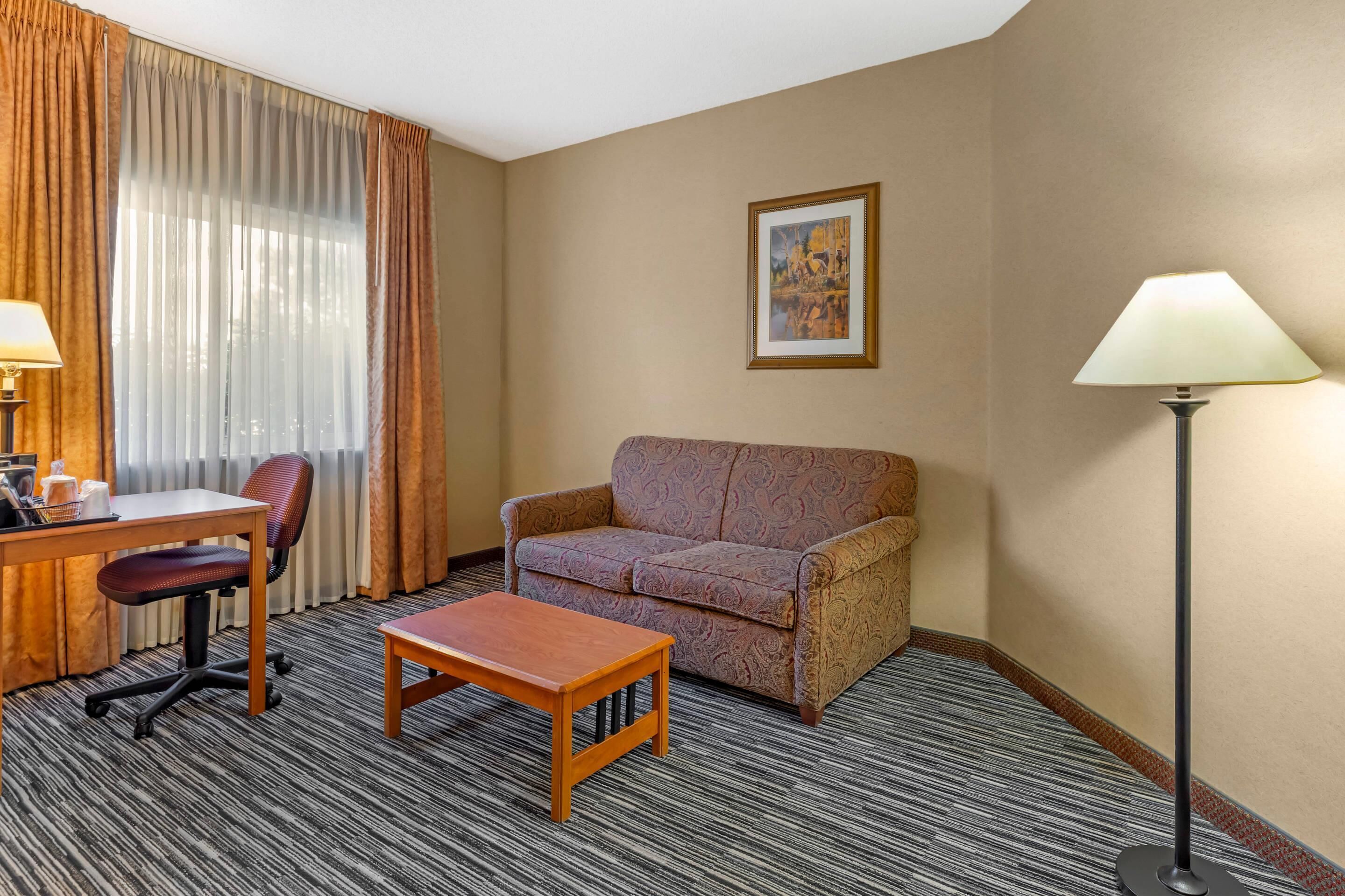 A hotel room with a sofa, a small table, a desk with a chair, a lamp, and a framed picture on the wall.