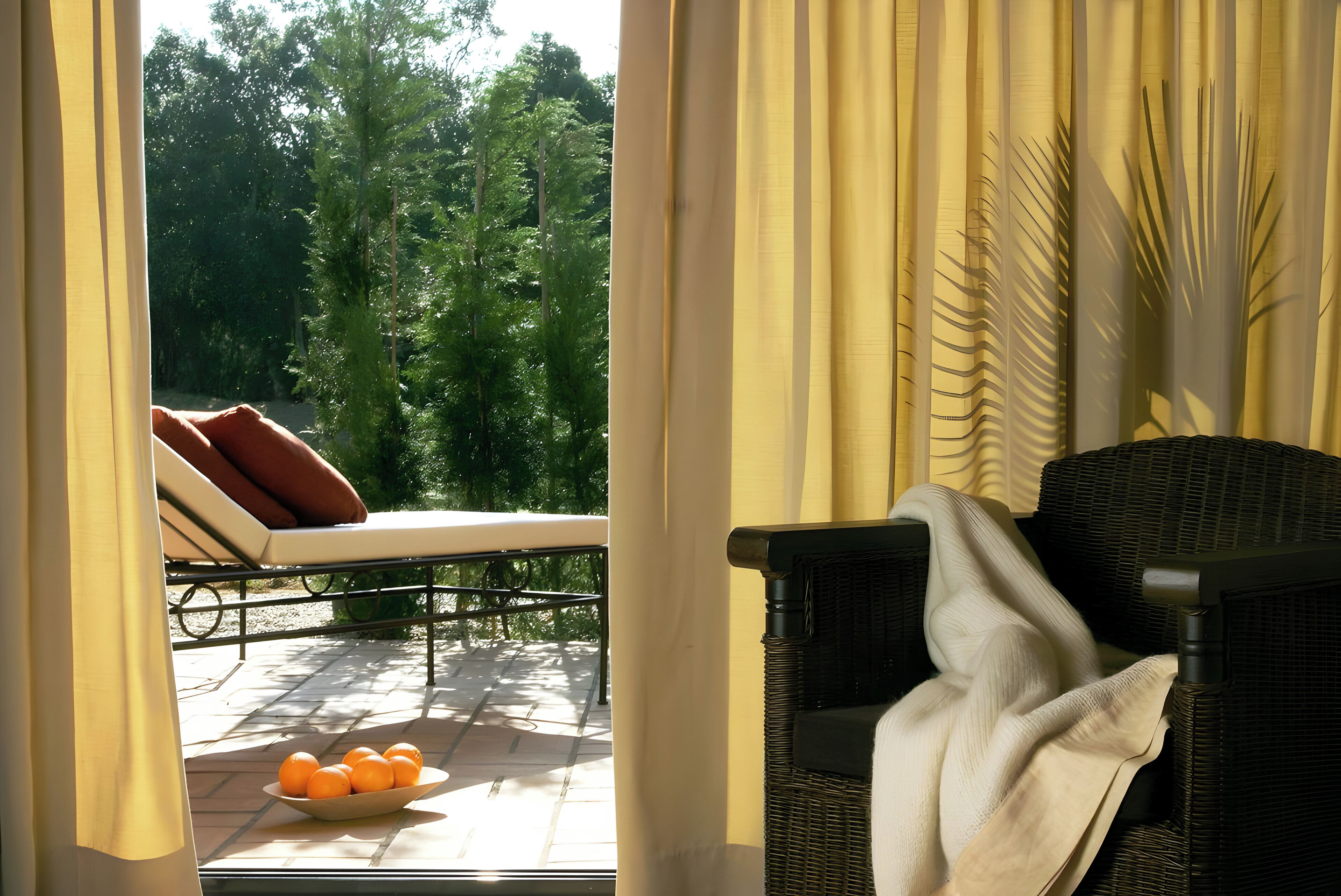 A room with a view of a garden, a wicker chair, a lounge chair with cushions, and a bowl of oranges on the windowsill.