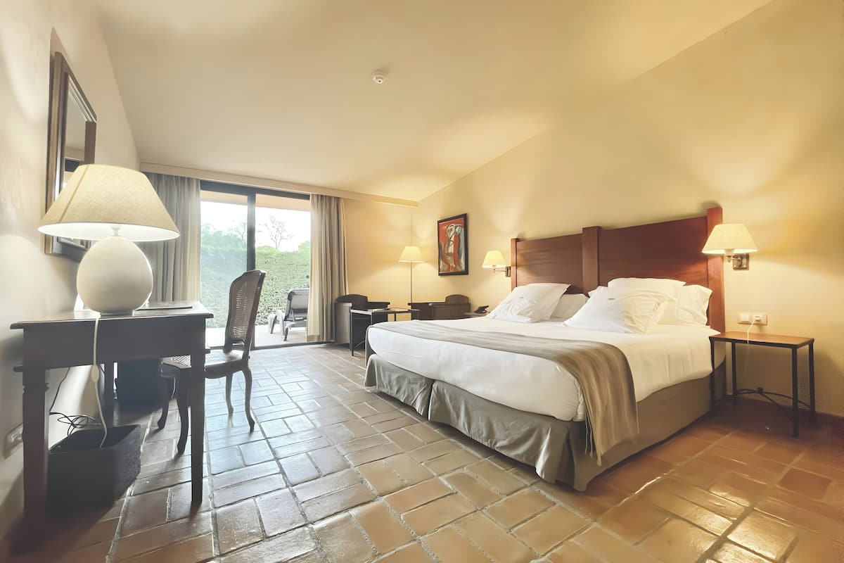 A hotel room with a bed, two bedside tables with lamps, a desk with a chair, a window with curtains, and a painting on the wall.