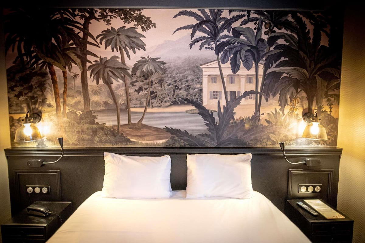 A hotel room with a large tropical mural, a bed with white linens, two bedside tables with lamps, and a nightstand with a phone and a remote control.