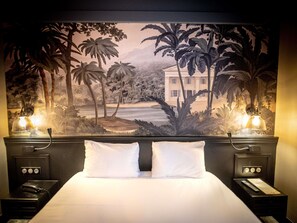 A hotel room with a large tropical mural, a bed with white linens, two bedside tables with lamps, and a nightstand with a phone and a remote control. - Mercure Rochefort La Corderie Royale (Rochefort)