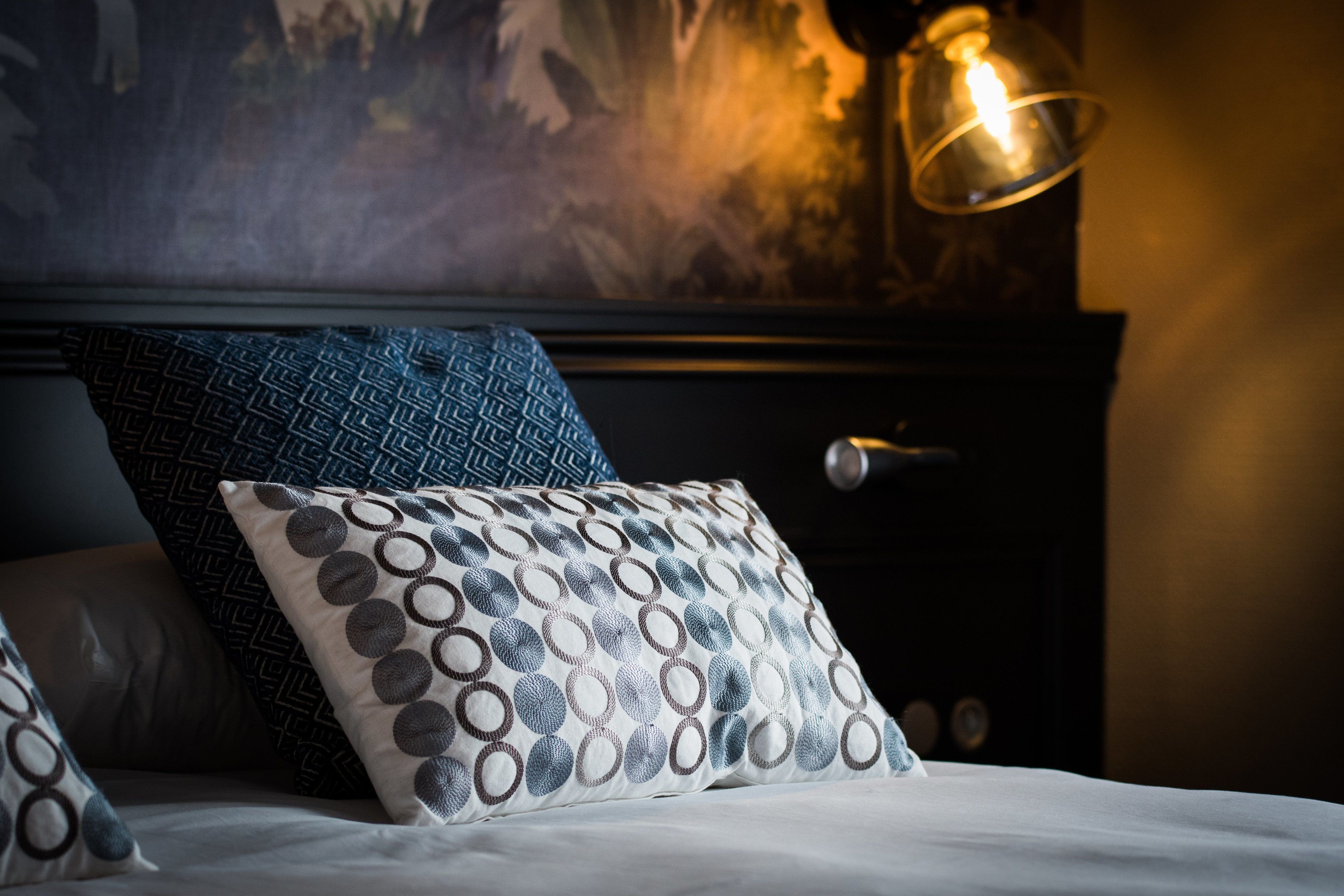 Una cama bien hecha con almohadas estampadas, cabecera oscura y lámpara fijada a la pared.