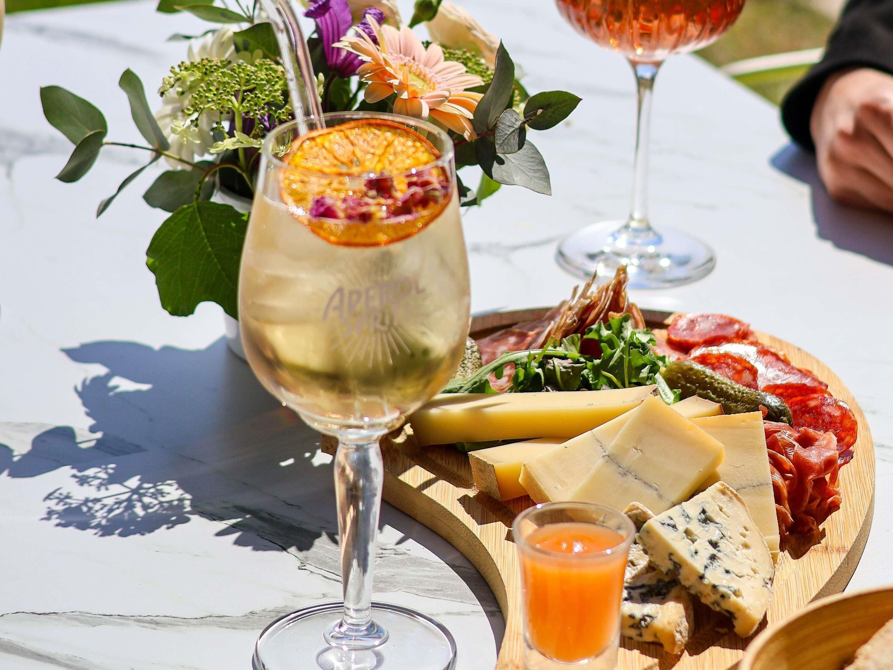 A table with a glass of white wine, a wooden board with various cheeses, cured meats, and a small glass of orange juice.