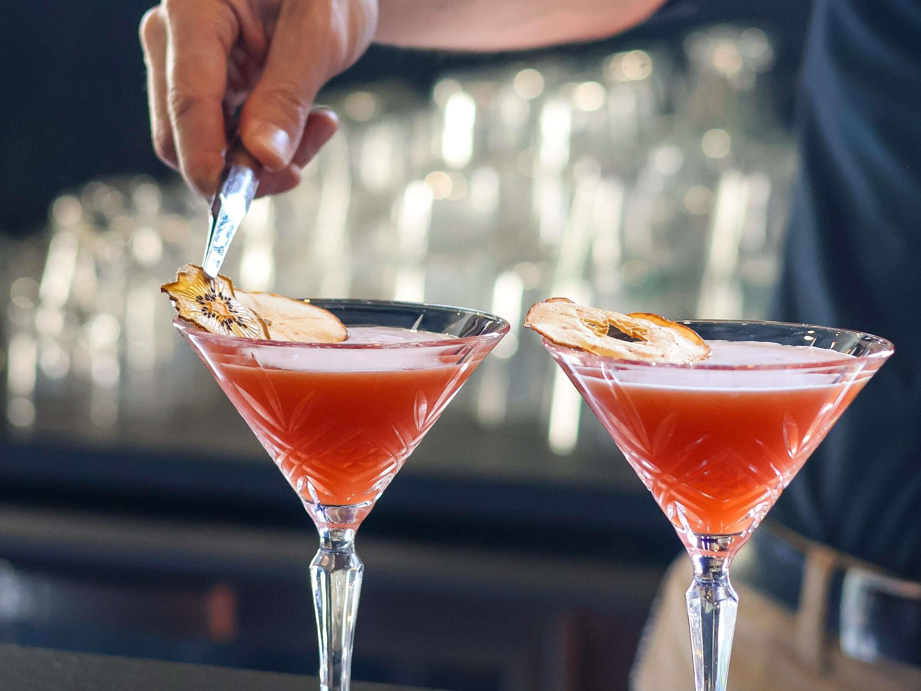 Dos copas de martini llenas de un brebaje rojo, adornadas con una rodaja de cáscara de naranja, sobre una barra.