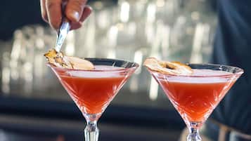 Dos copas de martini llenas de un brebaje rojo, adornadas con una rodaja de cáscara de naranja, sobre una barra.