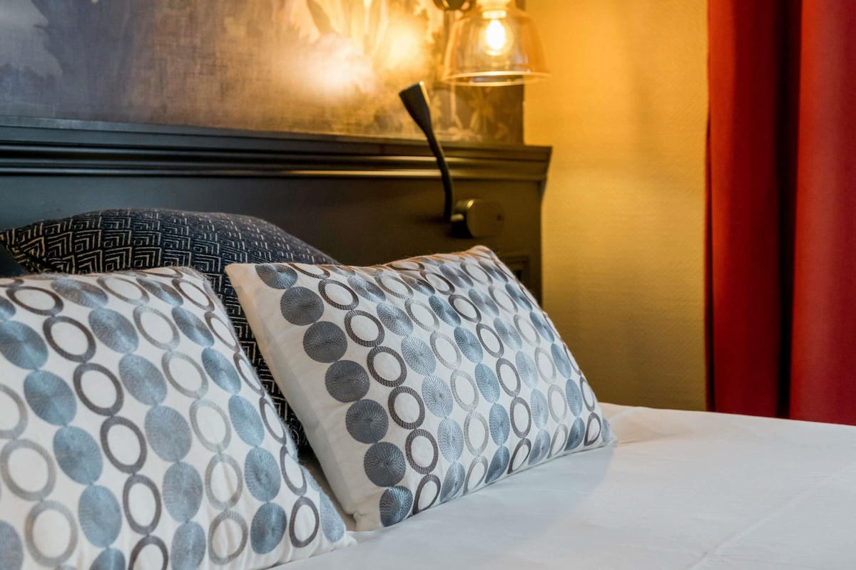 A bed with patterned pillows, a wall-mounted lamp, and a partial view of a painting.