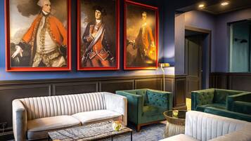 A modern hotel lobby with a white sofa, green armchairs, and three framed portraits on the wall.