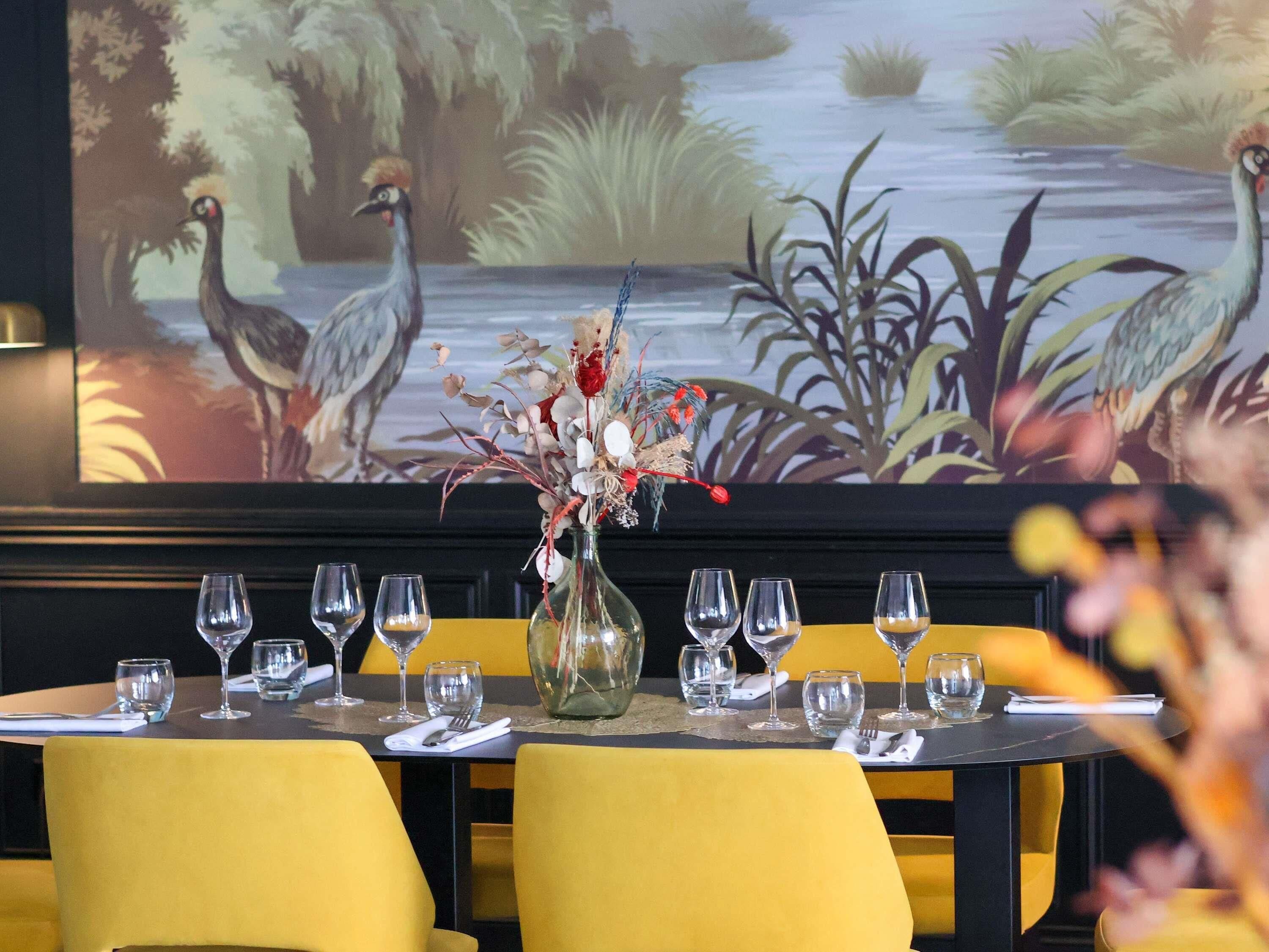 Una mesa de restaurante dispuesta con sillas amarillas, un jarrón con flores y un mural al fondo con aves y plantas.