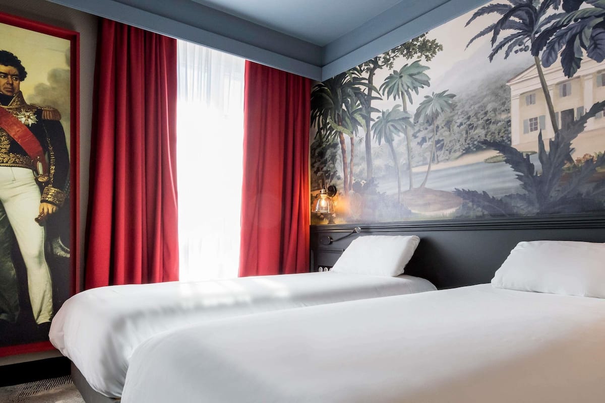 A hotel room with two beds, red curtains, a large portrait on the wall, and a mural depicting a tropical landscape.