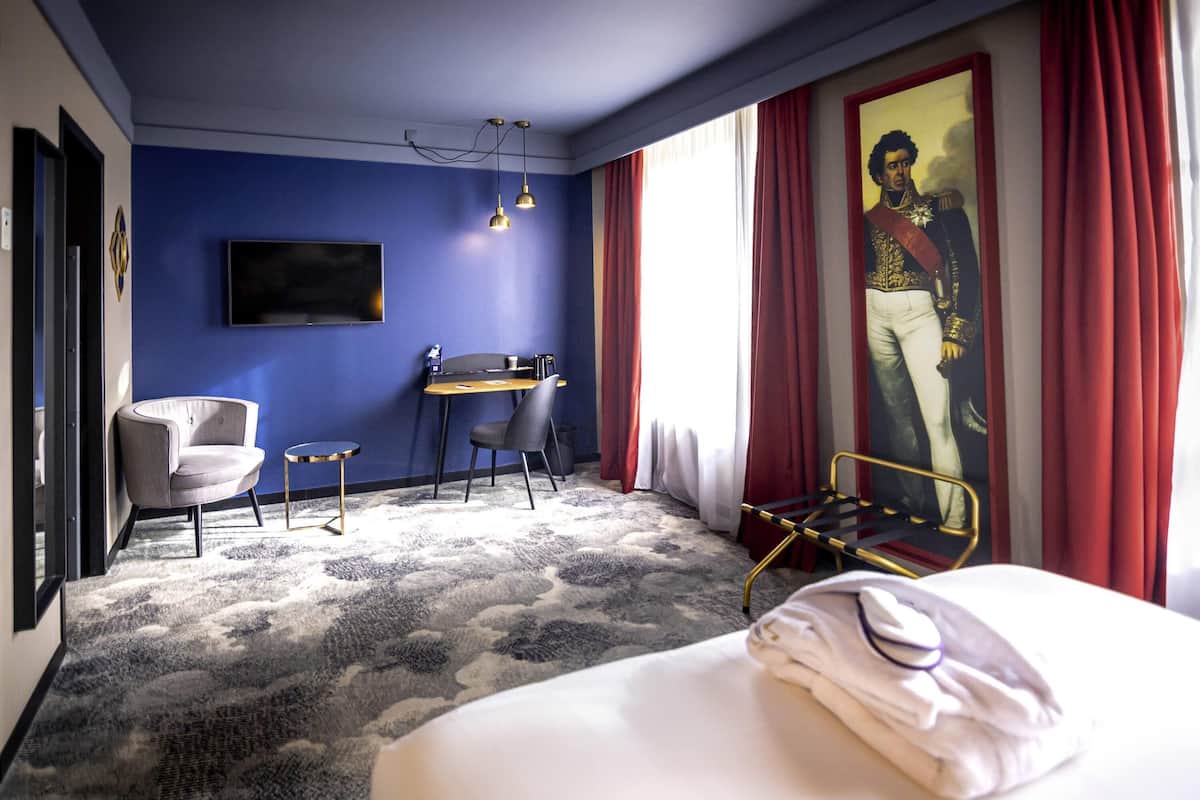 A hotel room with a bed, a desk, two chairs, a television, and a large window with red curtains.