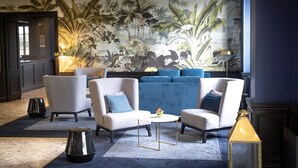 Un área de descanso moderna con un gran mural de plantas tropicales, un conjunto de sofás azules y una pequeña mesa redonda con lámpara.