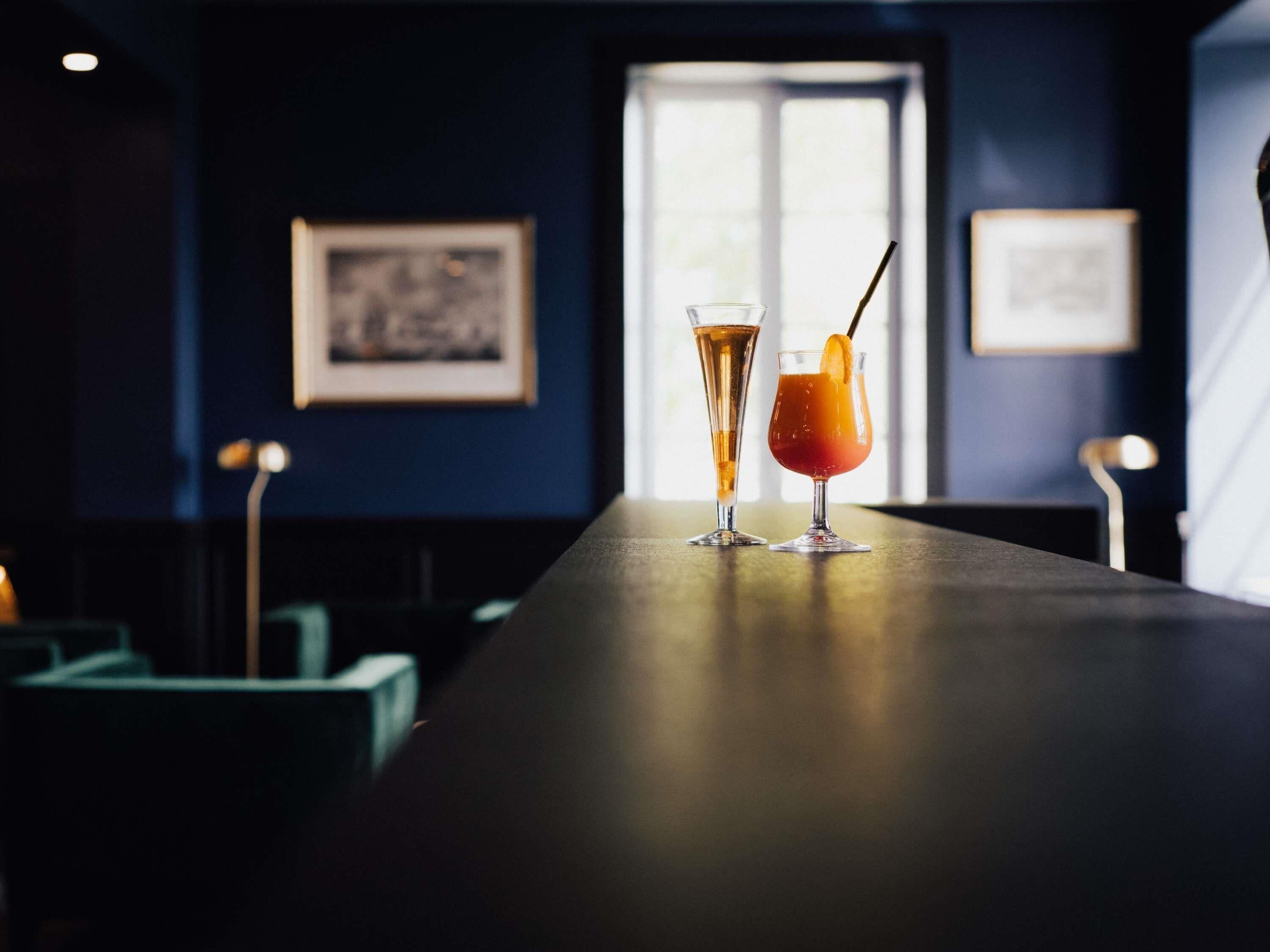 A bar with two drinks on a counter, framed pictures on the wall, and a window in the background.