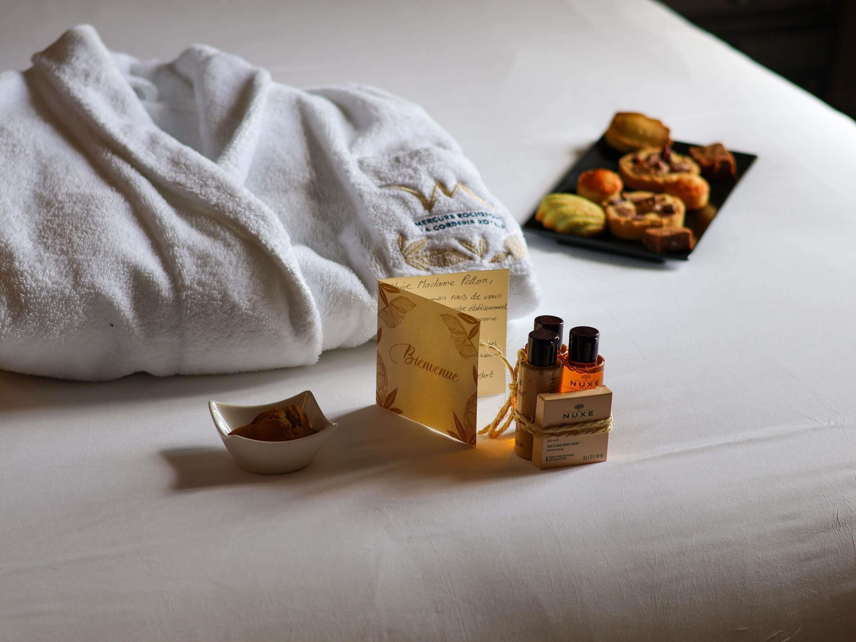 A neatly made bed with a white towel, a small bowl of cookies, and a box of Nuxe body soap.