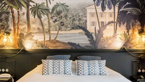 Habitación de hotel con una cama, dos almohadones estampados, una mesita de noche con teléfono y un mural de paisaje tropical.