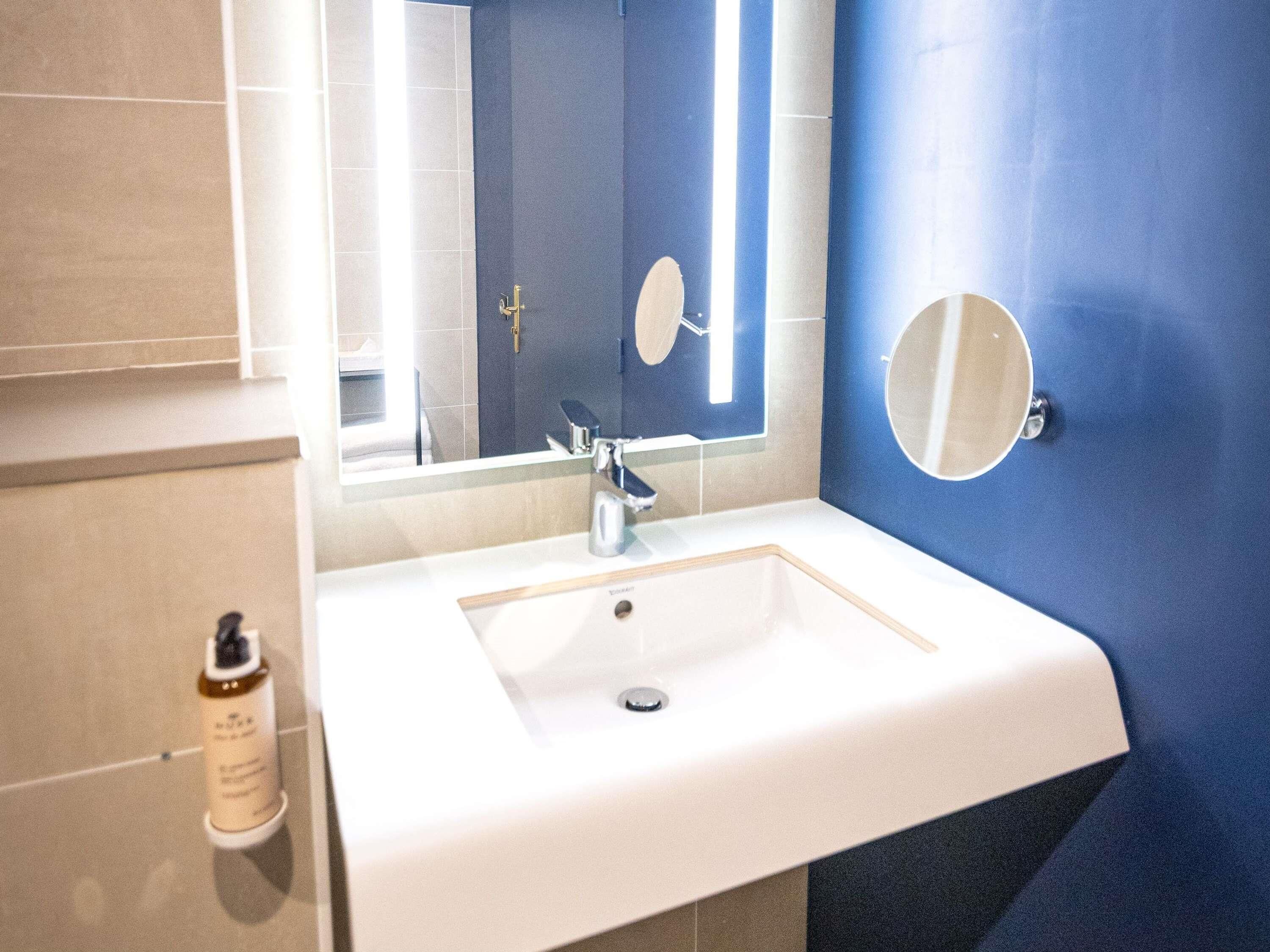 Un baño con un lavamanos blanco, un espejo y una botella de jabón para las manos.