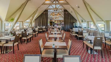 Un restaurant doté d’un plafond haut et de poutres apparentes, avec un tapis rouge à motifs et plusieurs tables à manger accompagnées de chaises.