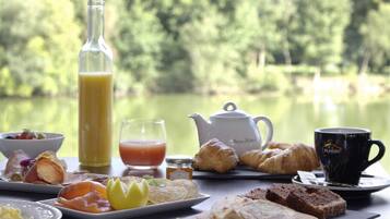 A breakfast table with croissants, pastries, a teapot, a mug, and a bottle of juice, set against a lake view.