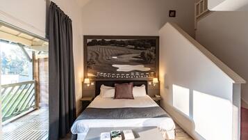 A bedroom with a large bed, a headboard with a landscape painting, bedside lamps, a wooden bench, and a view of a balcony.