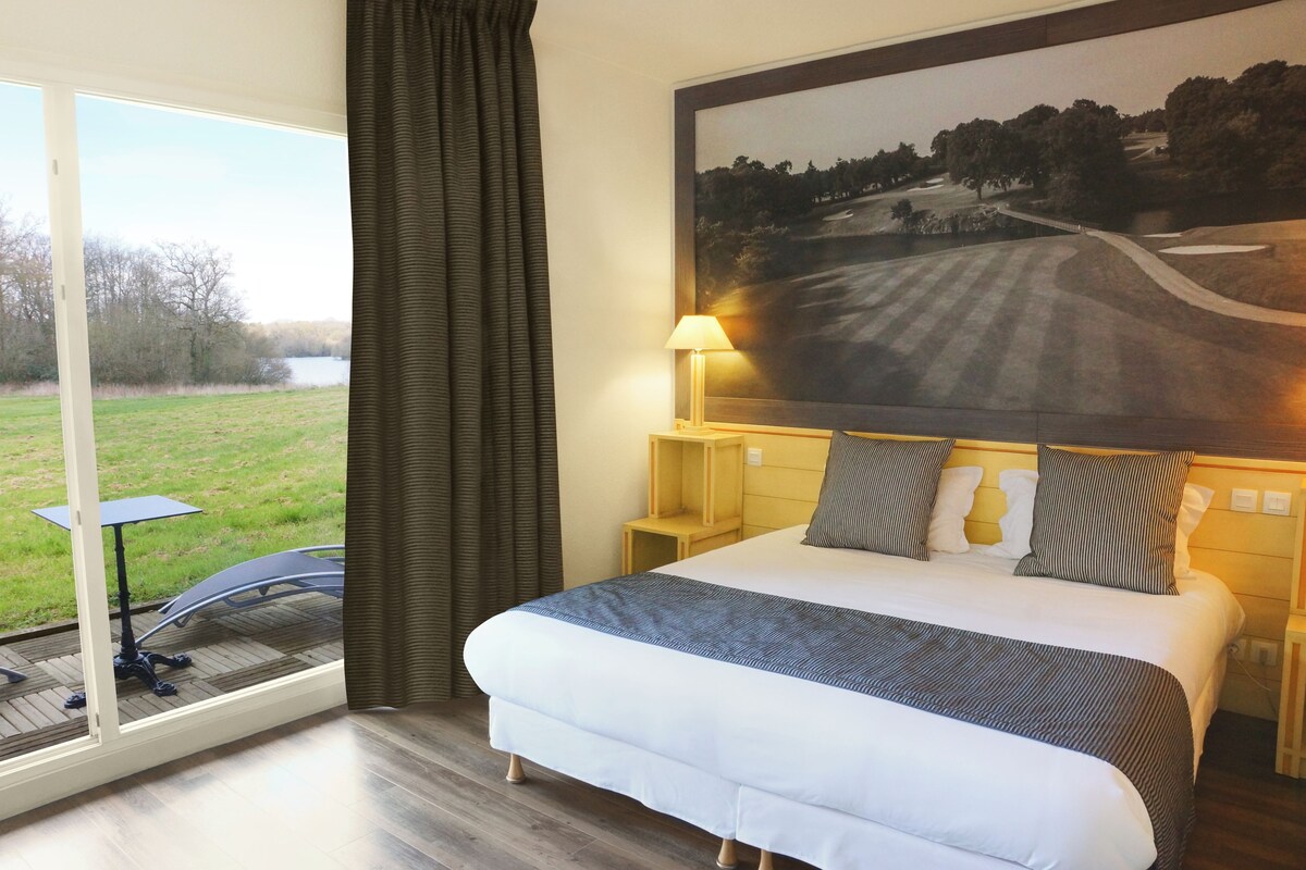 A bedroom with a large bed, a nightstand, a lamp, a window with curtains, and a view of a grassy area.