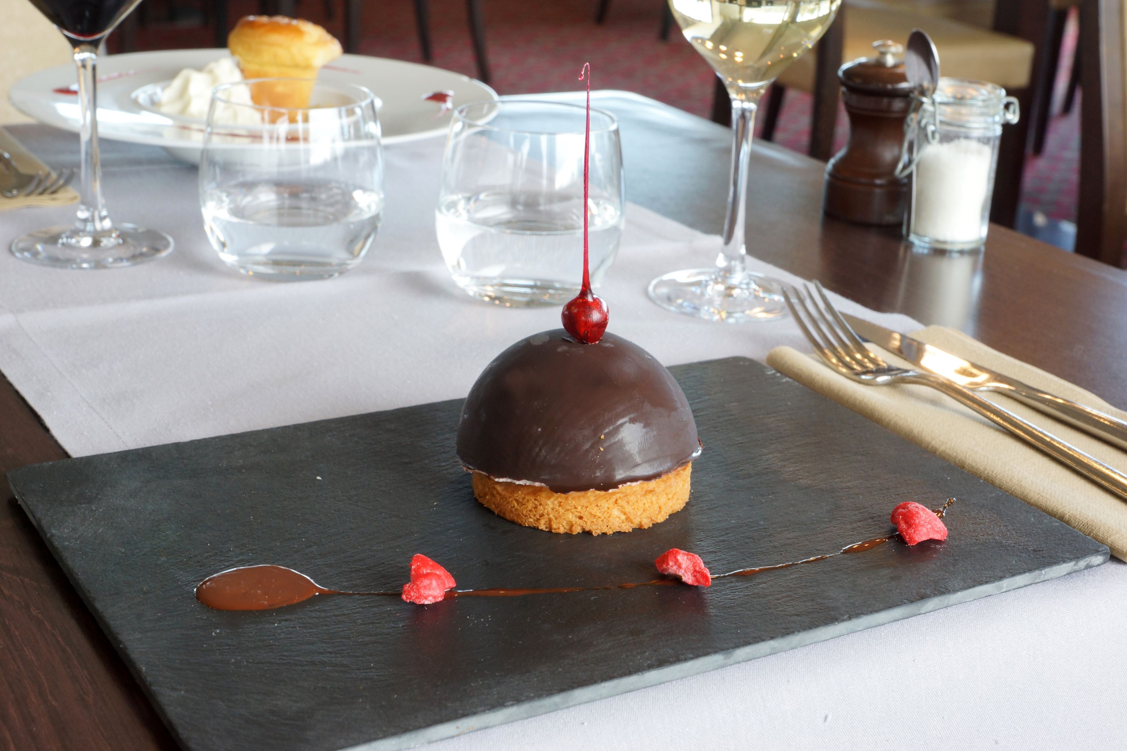 A dessert on a slate plate with a chocolate dome, a red berry, and a chocolate drizzle.