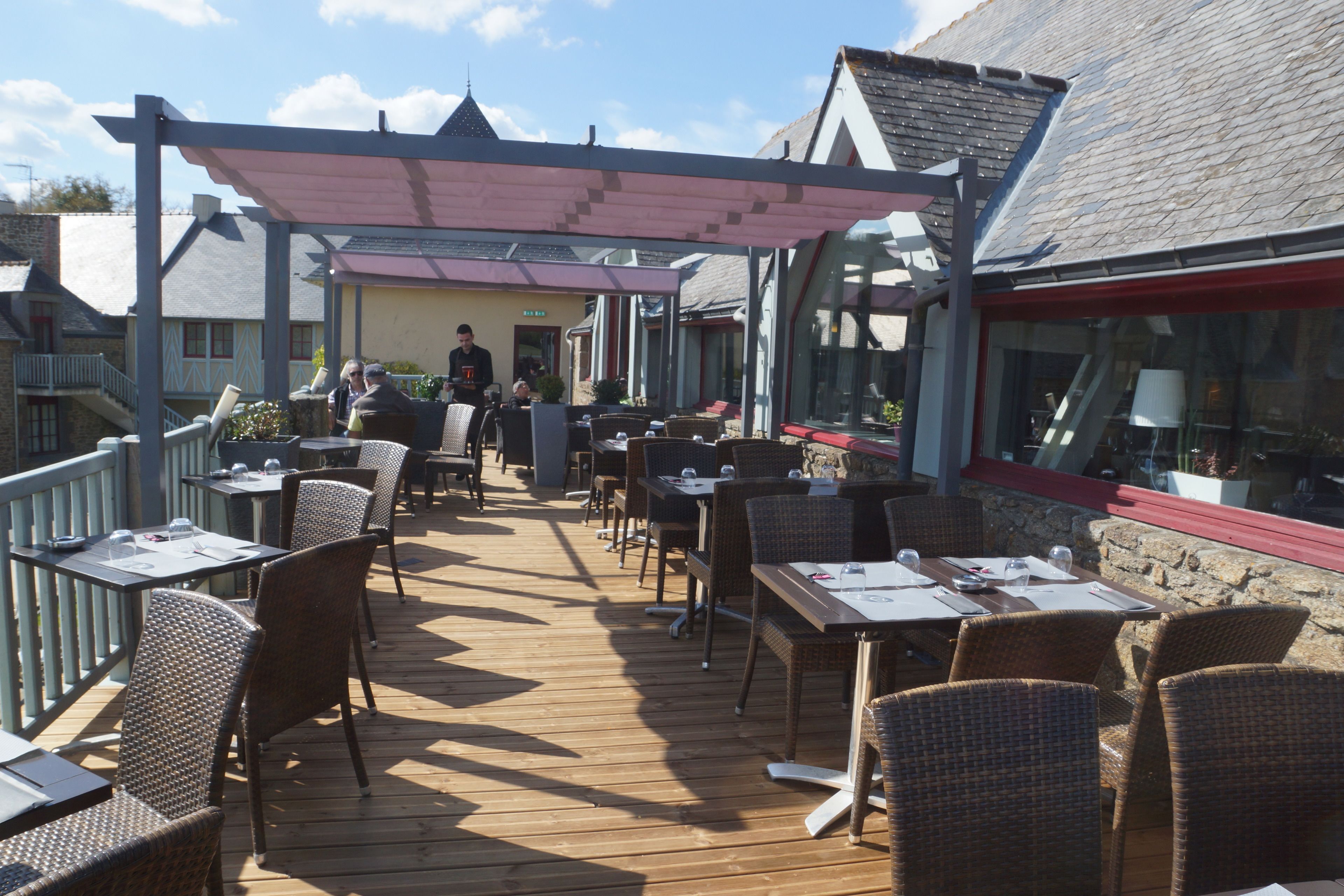 An outdoor patio with wooden tables and wicker chairs, a covered walkway, and a building with a sloped roof in the background.