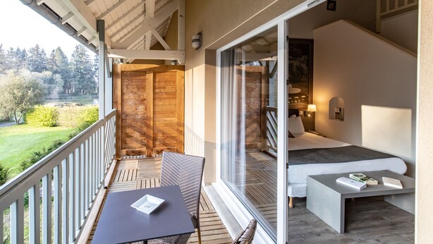 Een balkon met een houten tafel en stoelen, een bed met uitzicht op de omgeving, en een trap die naar een ander niveau leidt.