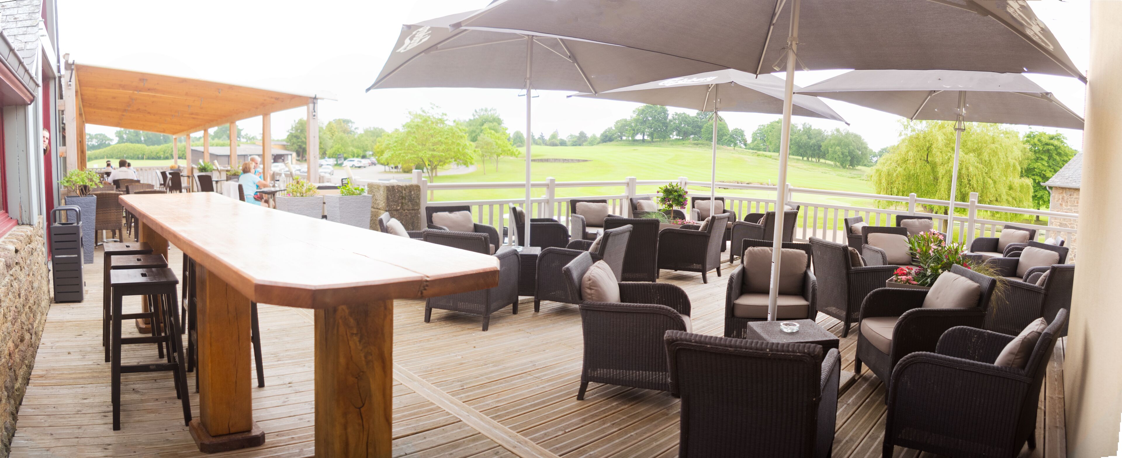 A covered outdoor seating area with a long wooden table and multiple chairs, overlooking a golf course.