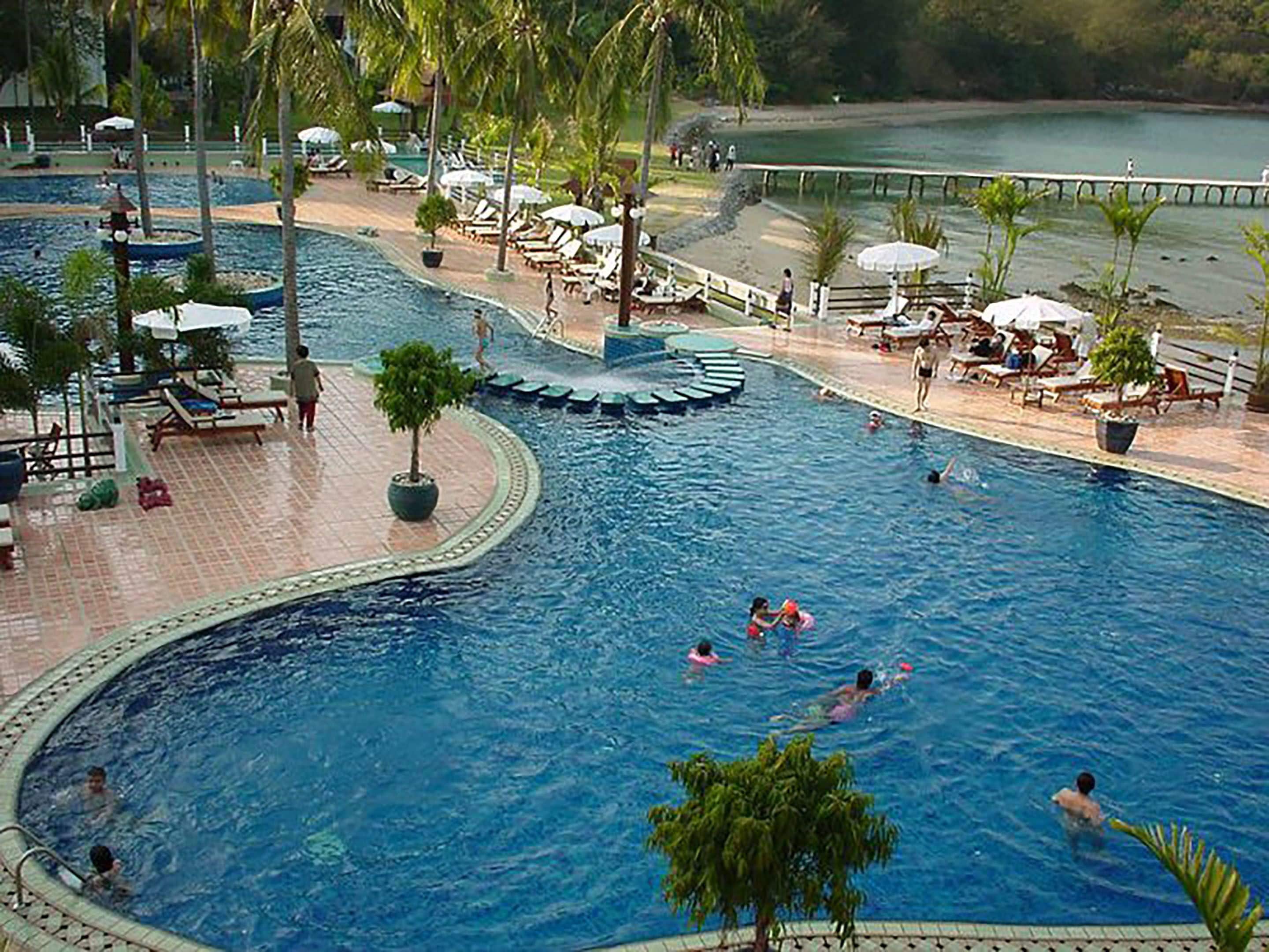 Una gran piscina al aire libre con múltiples carriles, rodeada de sillas de descanso y sombrillas, y vista a la playa y al agua en el fondo.