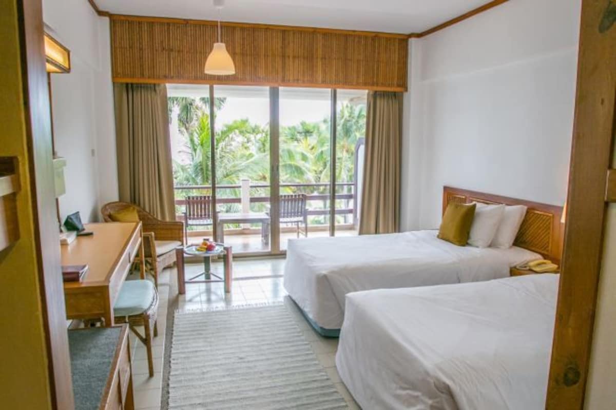 Habitación de hotel con dos camas, un escritorio y una silla. Tiene un balcón con vistas a palmeras.