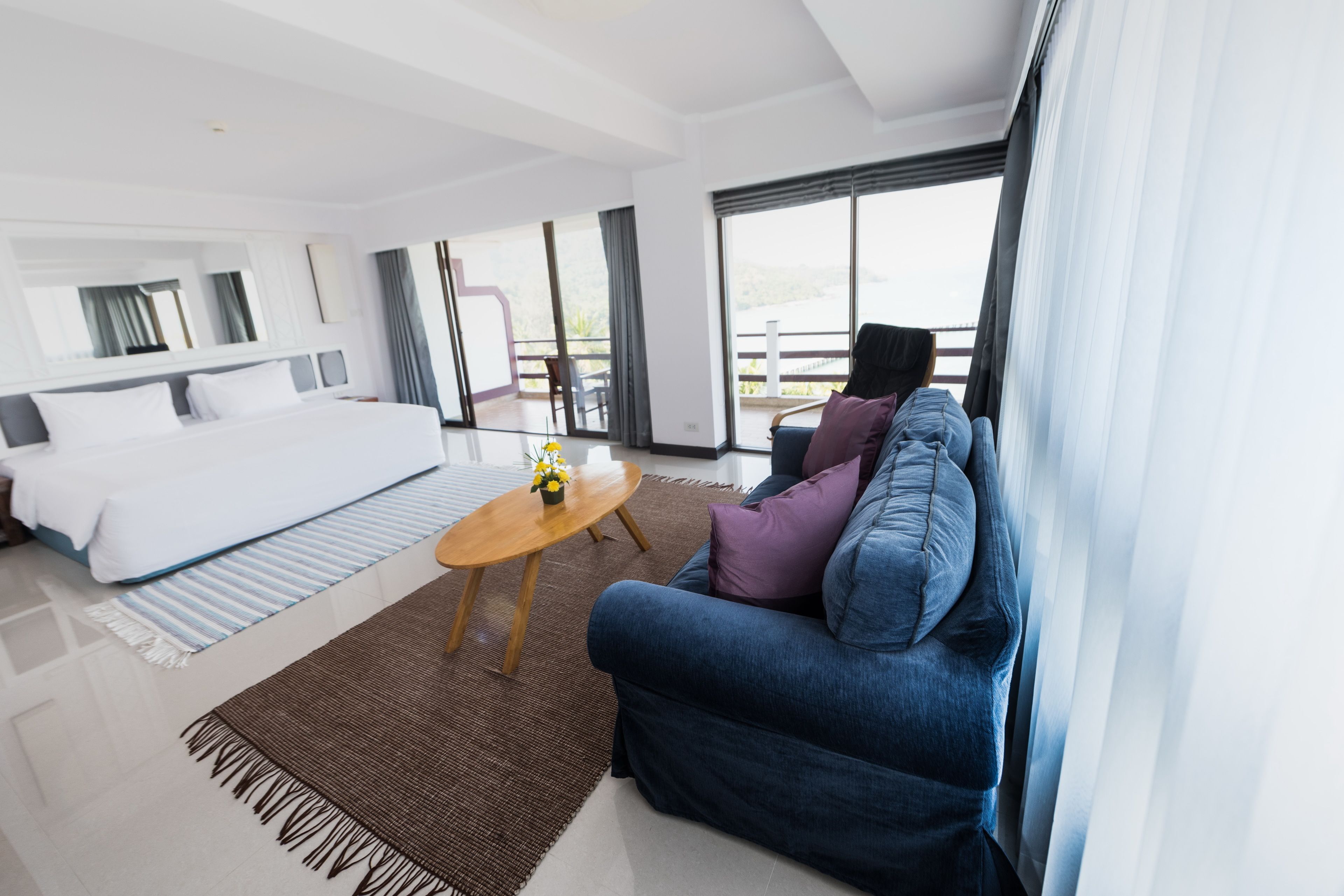 A modern hotel room with a large bed, a sofa, a small round table, and a balcony with a view.
