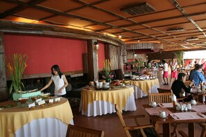 A restaurant with round tables covered in yellow tablecloths, wooden chairs, and a red wall.