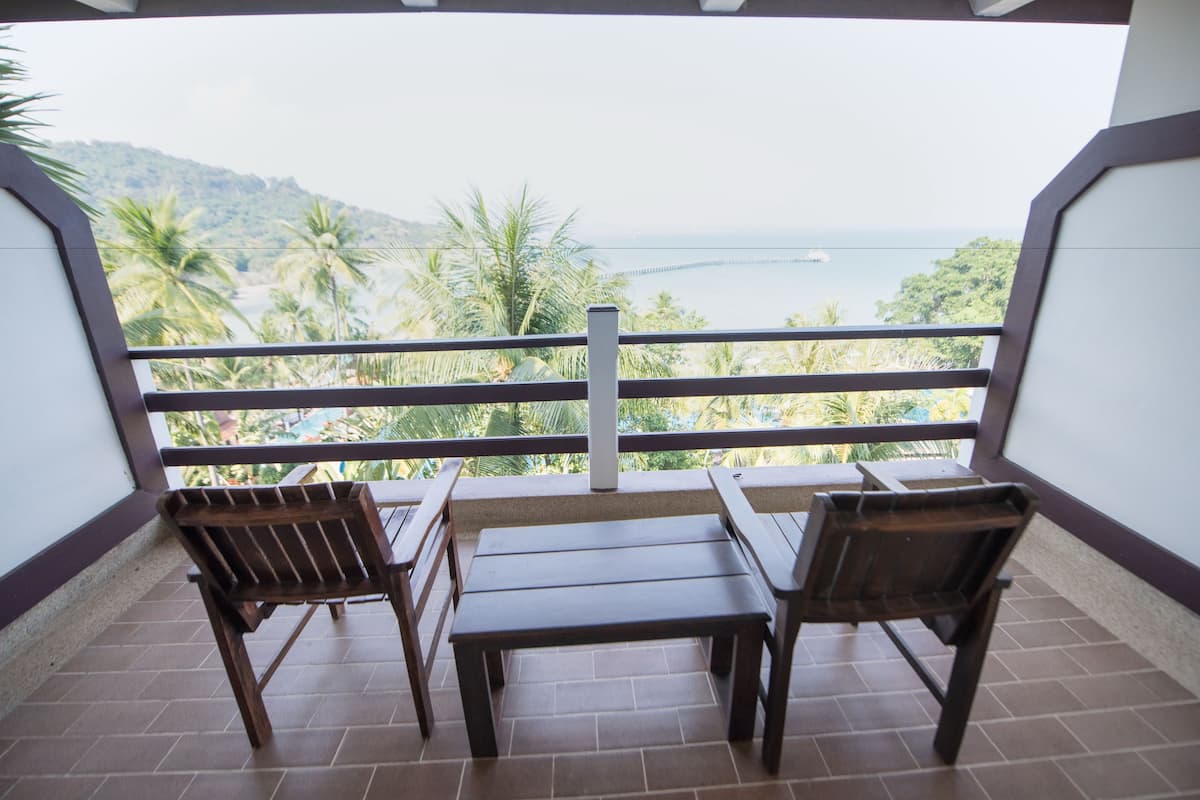 Un balcón con dos sillas de madera y una mesa, con vistas a la costa, palmeras y un puente.