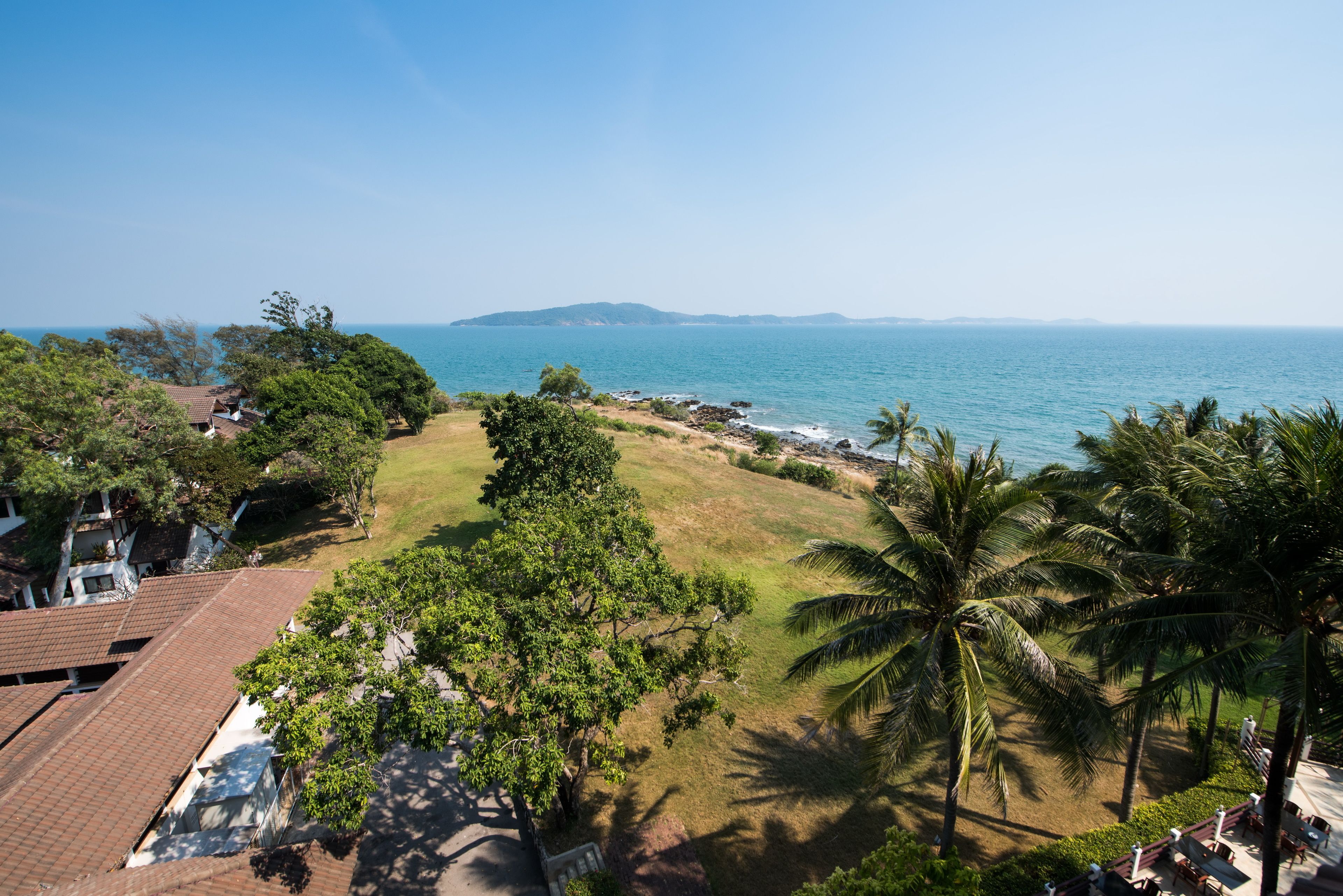 A coastal hotel with a view of the sea, palm trees, and a grassy area.