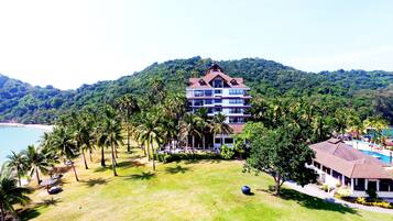 Un hotel de varios pisos con un patio central rodeado de palmeras, con vista a una playa y montañas al fondo.
